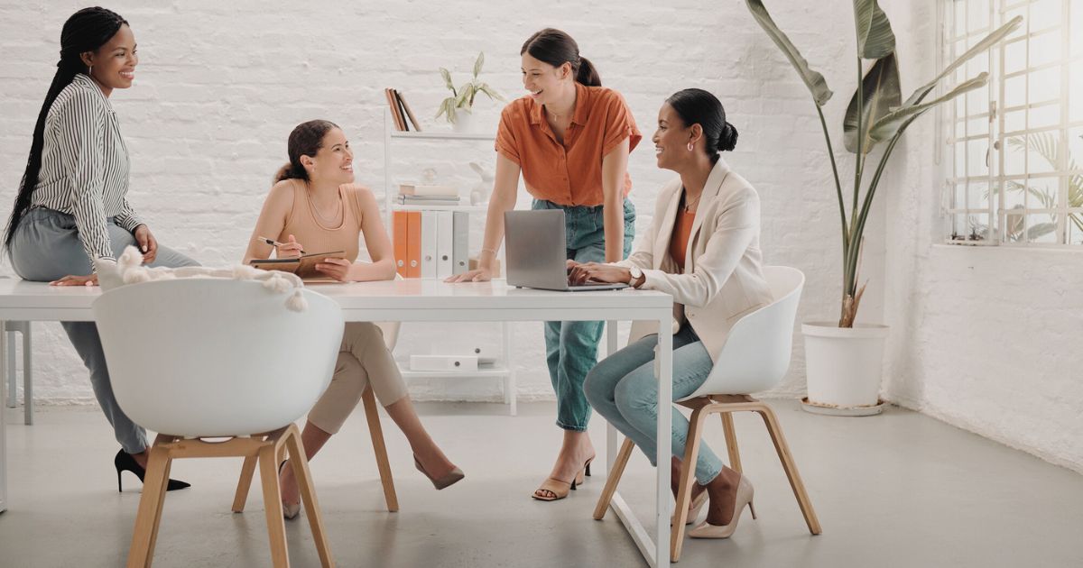 4 women sitting at a table in a work environment, laughing and talking.